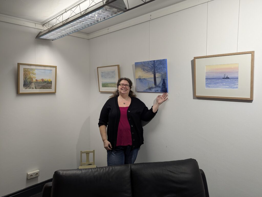 An einer Galerie-Wand hängen einige Bilder. Eine Dame, die Künstlerin, steht davor und präsentiert eines der Bilder.